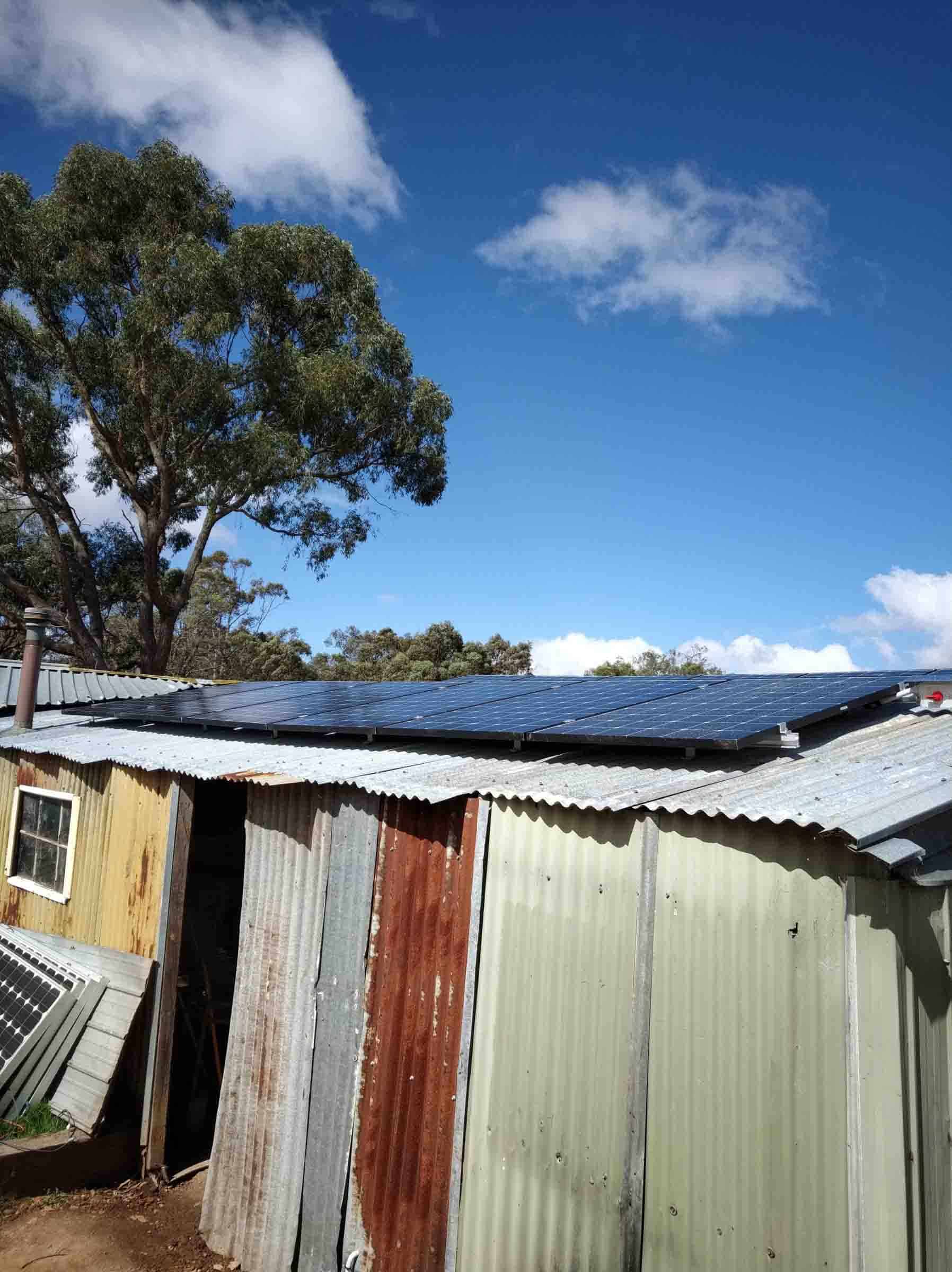 Solar panel array mounted on weathered corrugated iron shed roof with eucalyptus trees and blue sky in background
