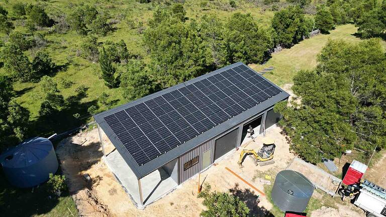 Large metal shed with complete rooftop solar panel array and construction equipment on rural property surrounded by trees