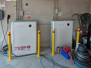 Two white SidEnergy battery units installed in garage with yellow safety bollards and electrical conduit connections