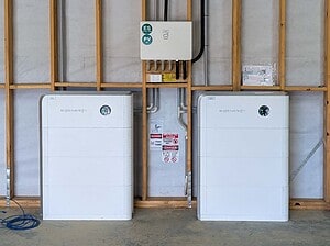 Two white Sidenergy battery units with solar inverter mounted above in wooden frame construction