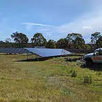 Large ground-mounted solar panel array in rural field with white 4WD vehicle and trees in background under blue sky