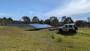 Large ground-mounted solar panel array in rural field with white 4WD vehicle and trees in background under blue sky