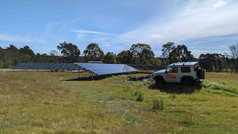 Large ground-mounted solar panel array in rural field with white 4WD vehicle and trees in background under blue sky