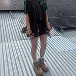 Female solar technician standing on corrugated metal roof with solar panels installed behind her, wearing work uniform