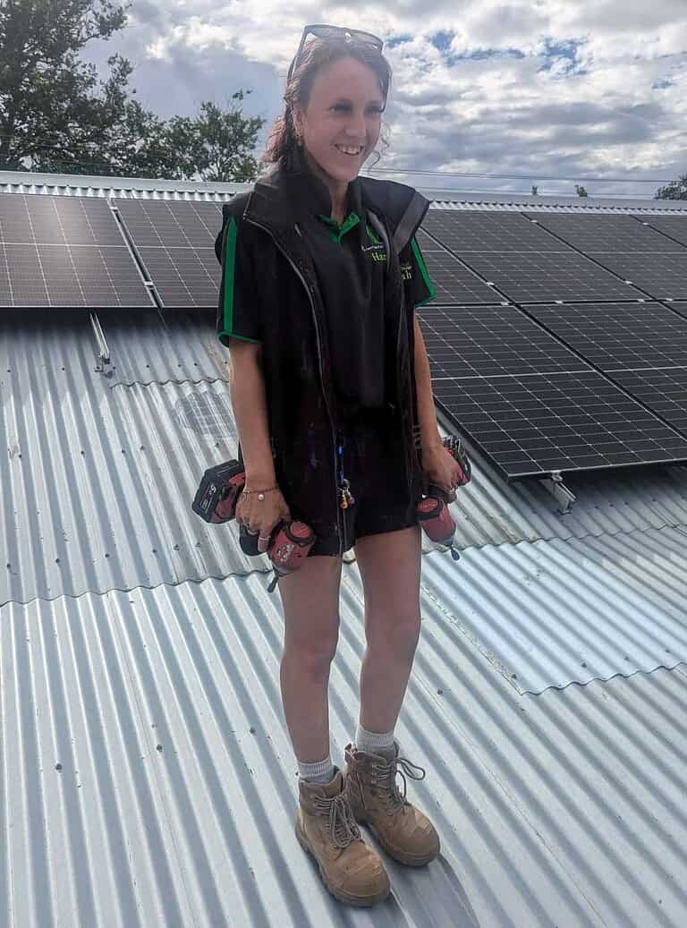 Female solar technician standing on corrugated metal roof with solar panels installed behind her, wearing work uniform