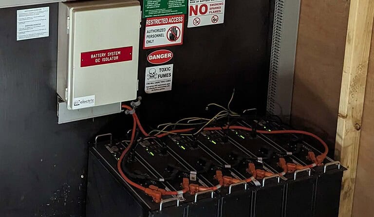 Black lithium battery modules with red DC cables and warning signs including restricted access and toxic fumes notices