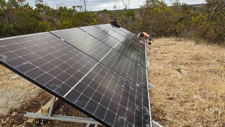Ground-mounted solar panel array being installed in rural bushland setting with technician working on connections