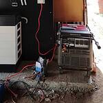 BYD battery storage unit connected to Honda generator via red charging cables in concrete-floored garage or shed