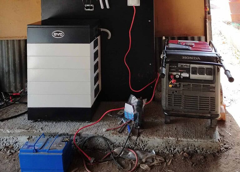 BYD battery storage unit connected to Honda generator via red charging cables in concrete-floored garage or shed
