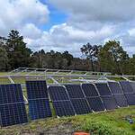 Multiple solar panels mounted on metal framework in rural field with trees in background and installer working