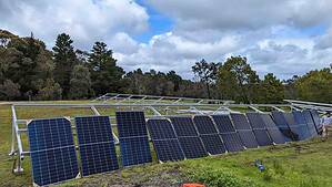 Multiple solar panels mounted on metal framework in rural field with trees in background and installer working