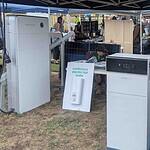 Solar equipment display under tent showing Pylontech battery system, air conditioning unit, and continuous electric hot water setup