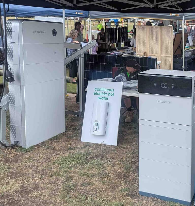 Solar equipment display under tent showing Pylontech battery system, air conditioning unit, and continuous electric hot water setup