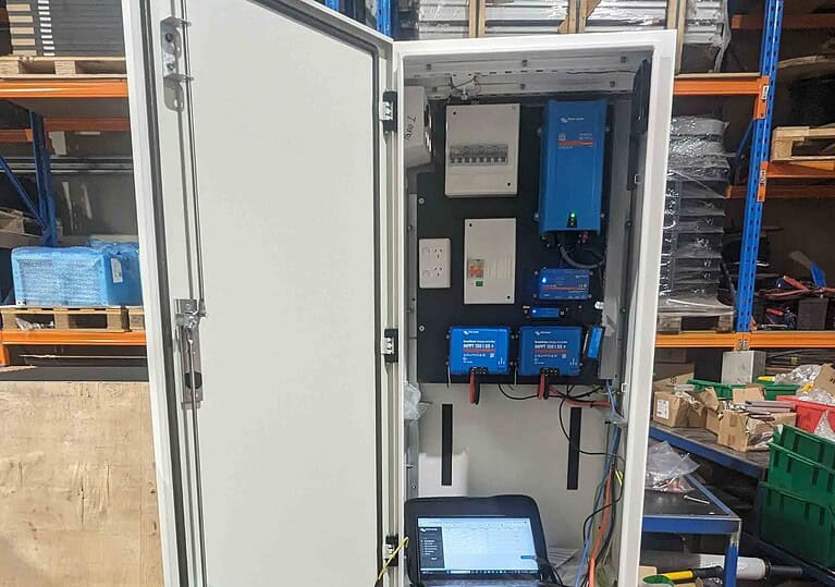 Open electrical cabinet showing Victron inverters, MPPT controllers, and monitoring equipment with laptop for configuration