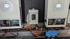 Two white Deye inverters with digital displays mounted above electrical switchboard with orange and black cables