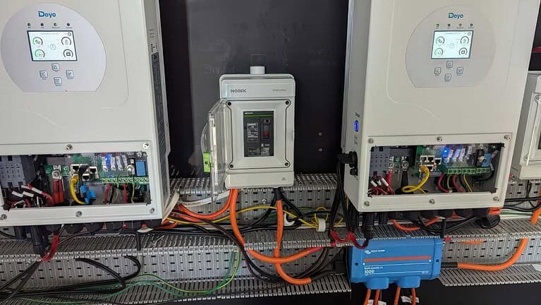Two white Deye inverters with digital displays mounted above electrical switchboard with orange and black cables