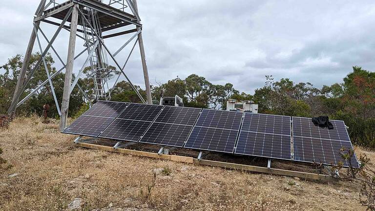 Ground-mounted solar panel array installed near telecommunications tower in rural Australian bushland setting