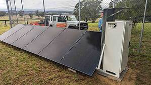 Ground-mounted solar panel array with white battery storage unit behind security fencing in rural Victorian setting