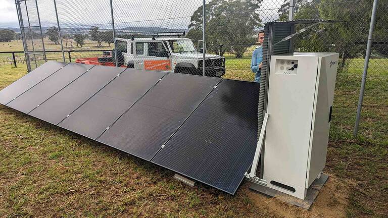 Ground-mounted solar panel array with white battery storage unit behind security fencing in rural Victorian setting