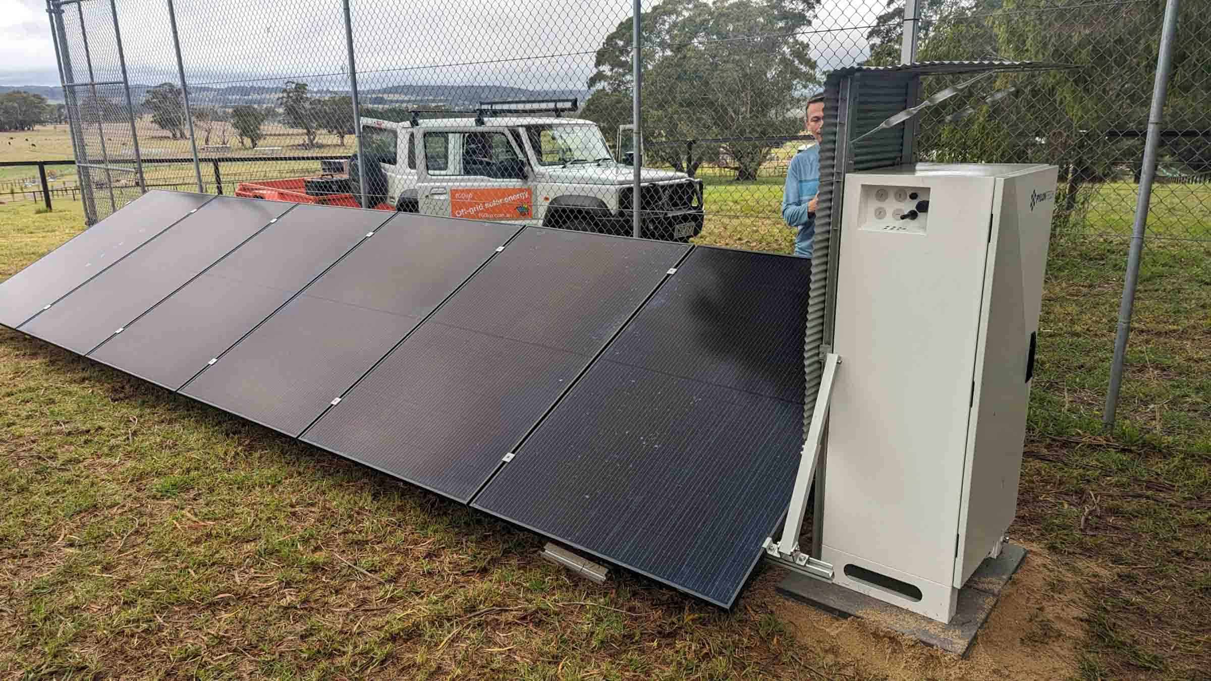 Ground-mounted solar panel array with white battery storage unit behind security fencing in rural Victorian setting