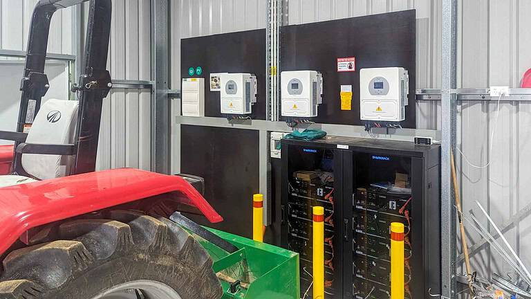 Black server rack containing stacked lithium batteries with three wall-mounted inverters above in Ballarat off-grid installation