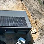 Aerial view of metal shed with solar panels covering most of the roof, located on cleared rural land with construction equipment