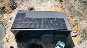 Aerial view of metal shed with solar panels covering most of the roof, located on cleared rural land with construction equipment