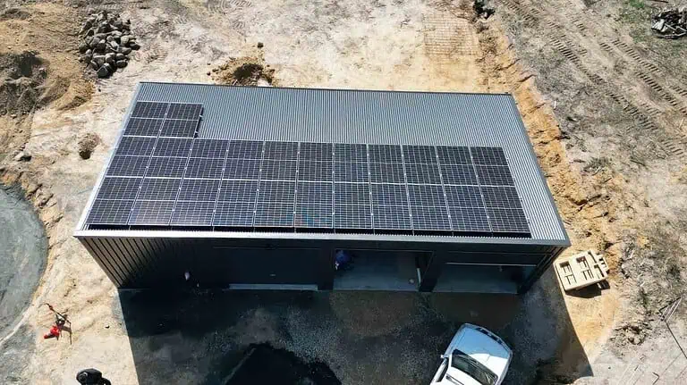 Aerial view of metal shed with solar panels covering most of the roof, located on cleared rural land with construction equipment