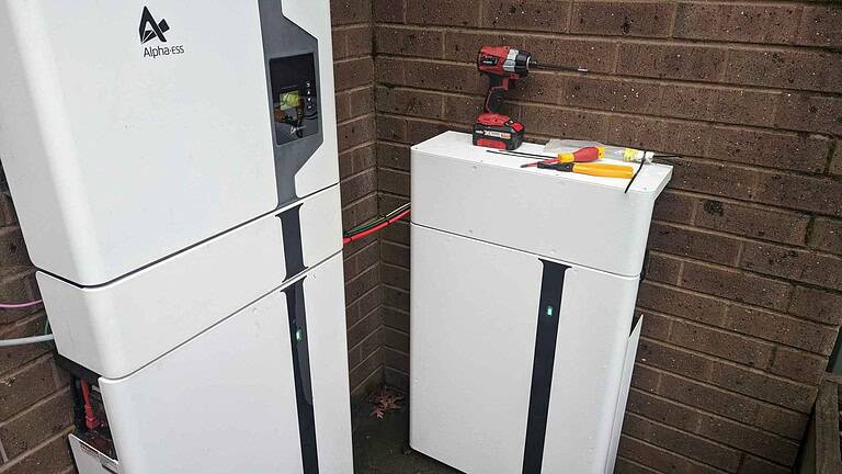 White AlphaESS battery storage units stacked against brick wall with red drill and tools on top during installation