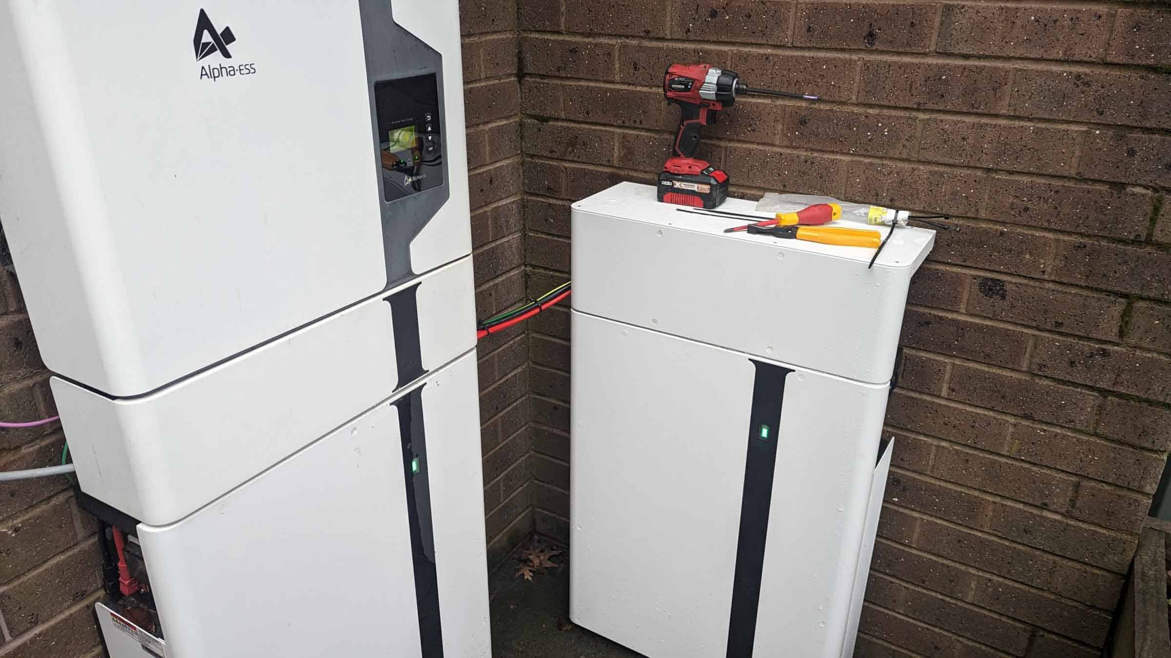White AlphaESS battery storage units stacked against brick wall with red drill and tools on top during installation