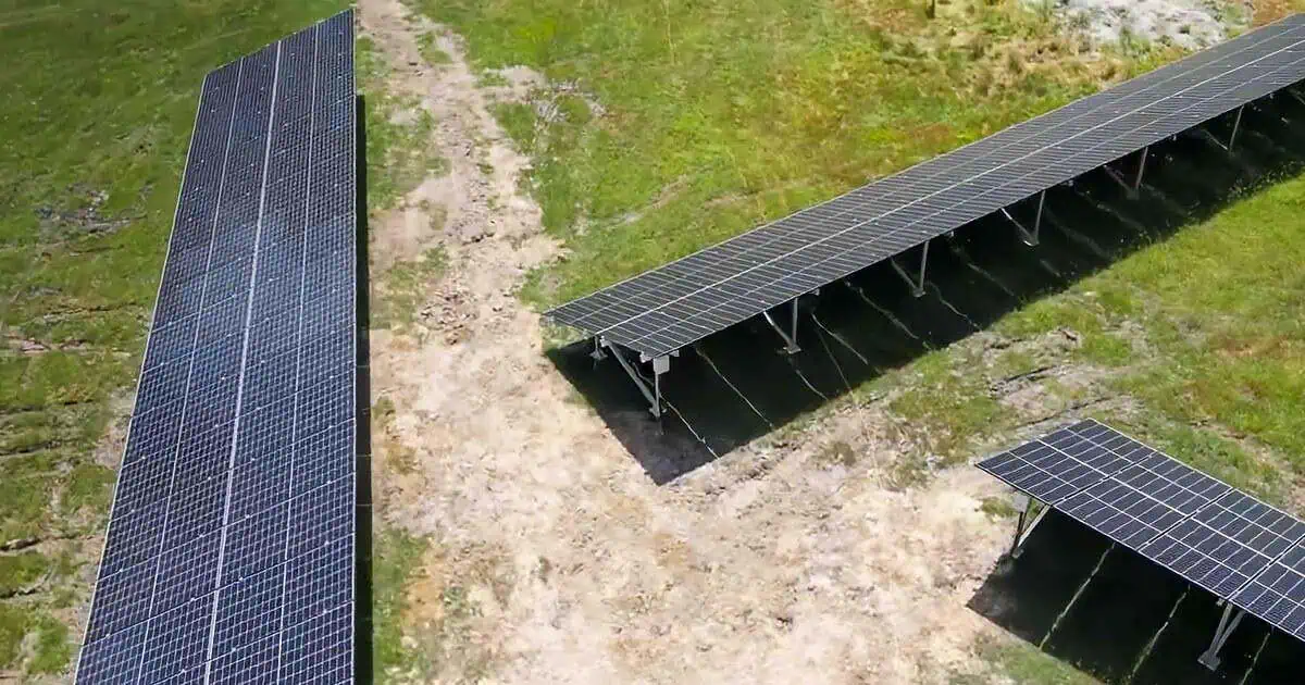 Aerial view of solar panel arrays installed on ground-mounted structures in a rural, grassy area with dirt paths and rocky outcrops